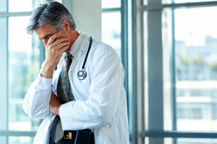 Medico con camice bianco e stetoscopio si copre il volto con una mano, manifestando stanchezza e stress lavorativo.